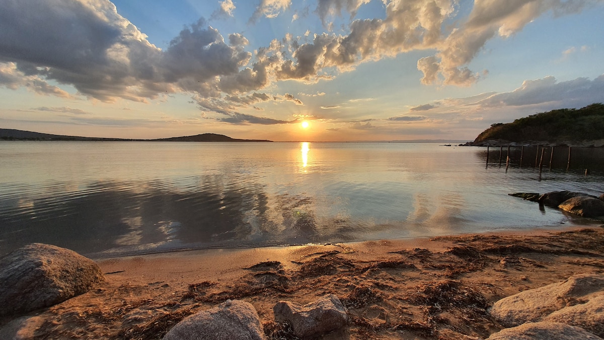 A serene beach scene is captured at sunset, featuring calm waters reflecting the orange and pink hues of the sky. The shoreline is framed by smooth stones and gentle waves, providing a peaceful backdrop for relaxation.