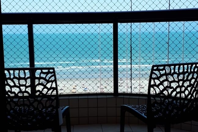 A balcony view is presented through a protective netting, showcasing the expansive sea and sandy beach below. Two chairs are positioned for relaxation, emphasizing the serene coastal setting. The clear blue water reflects the sky, creating a calming visual experience.