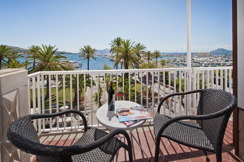 A small terrace is equipped with two black woven chairs and a round table. A bottle of wine and magazines are placed on the table. Palm trees and the marina are visible in the background, creating a vibrant coastal atmosphere.