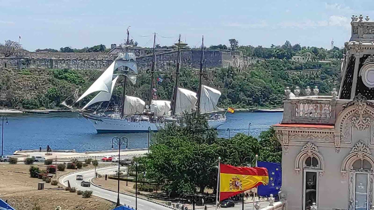 A sailing ship with white sails is anchored in the bay, accompanied by lush greenery along the shore. Historic structures are visible in the background against a clear blue sky. Nearby, flags flutter gently in the breeze, adding a touch of color to the tranquil waterfront.