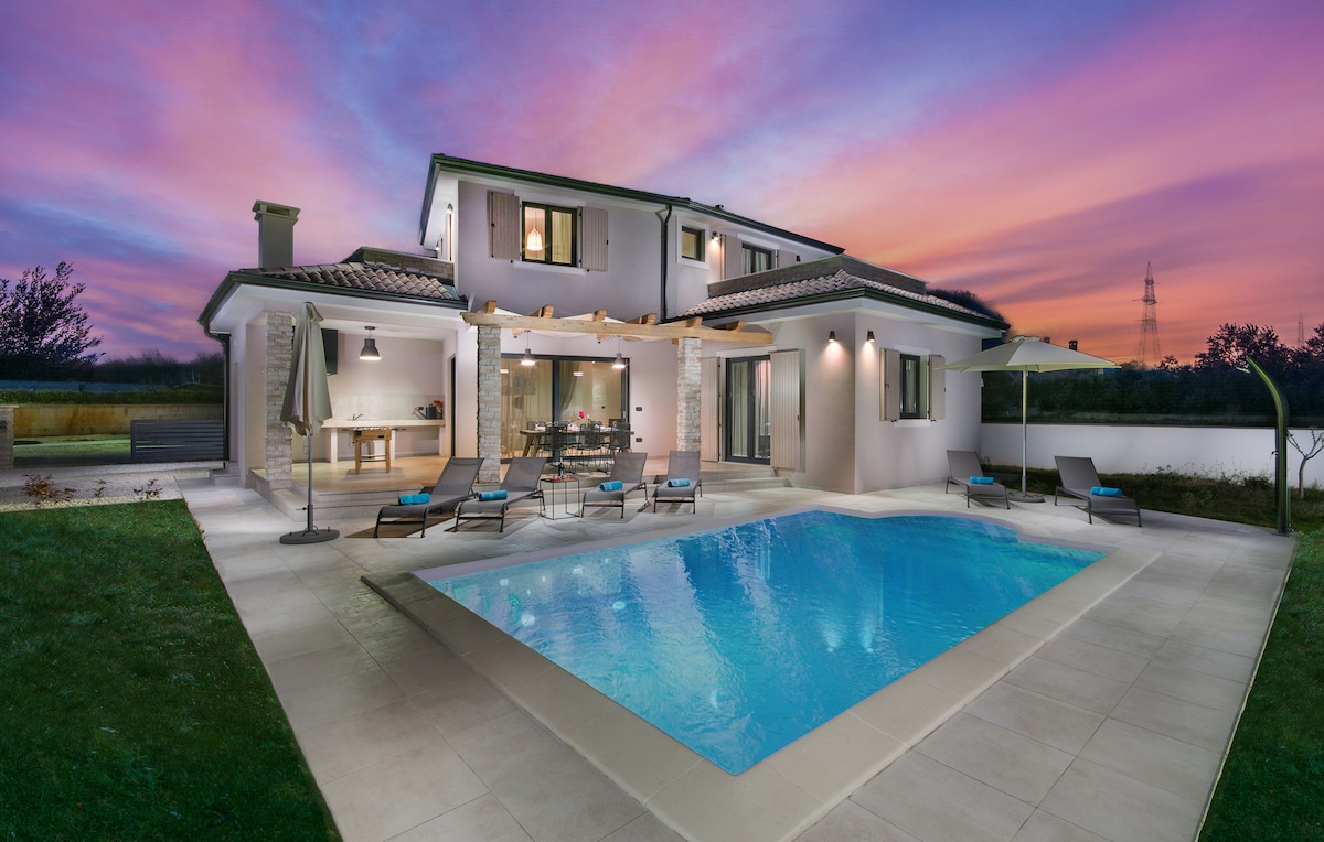A modern villa is depicted at dusk, showcasing a clear pool surrounded by loungers. A covered terrace leads to a spacious dining area. The two-story structure features large windows and a warm color palette, with a colorful sky creating a serene backdrop.