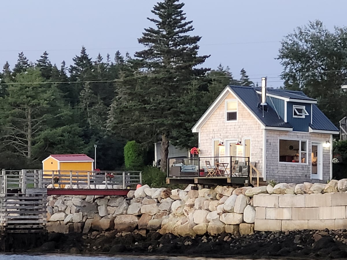 A charming coastal retreat is depicted, featuring a light-colored shingle exterior and a pitched metal roof. A wooden deck is shown, surrounded by lush greenery and rocky shoreline. Brightly colored storage shed stands nearby, enhancing the picturesque seaside landscape.