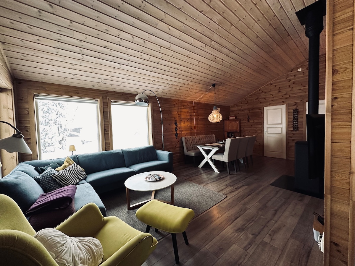 The spacious living area features a sectional sofa in a calming blue hue, complemented by a light wooden ceiling. A dining table with chairs is positioned near the window, while a cozy armchair in vibrant yellow adds a pop of color. A modern lighting fixture hangs above the dining space.