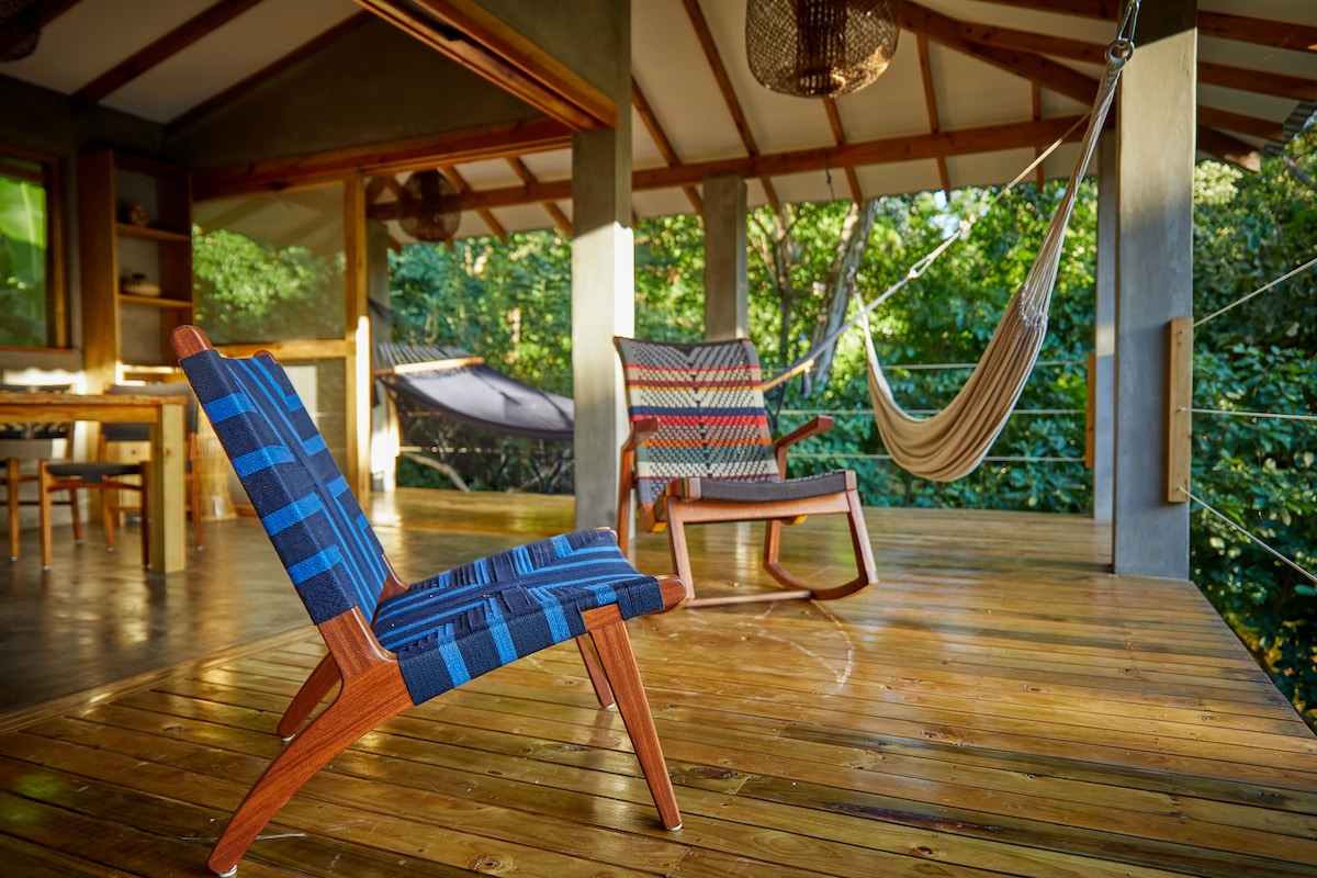 A spacious outdoor terrace features wooden flooring and comfortable seating options, including two chairs and a hammock. Lush greenery surrounds the space, providing a tranquil jungle atmosphere. Natural light fills the area through large openings, enhancing the seamless connection with nature.