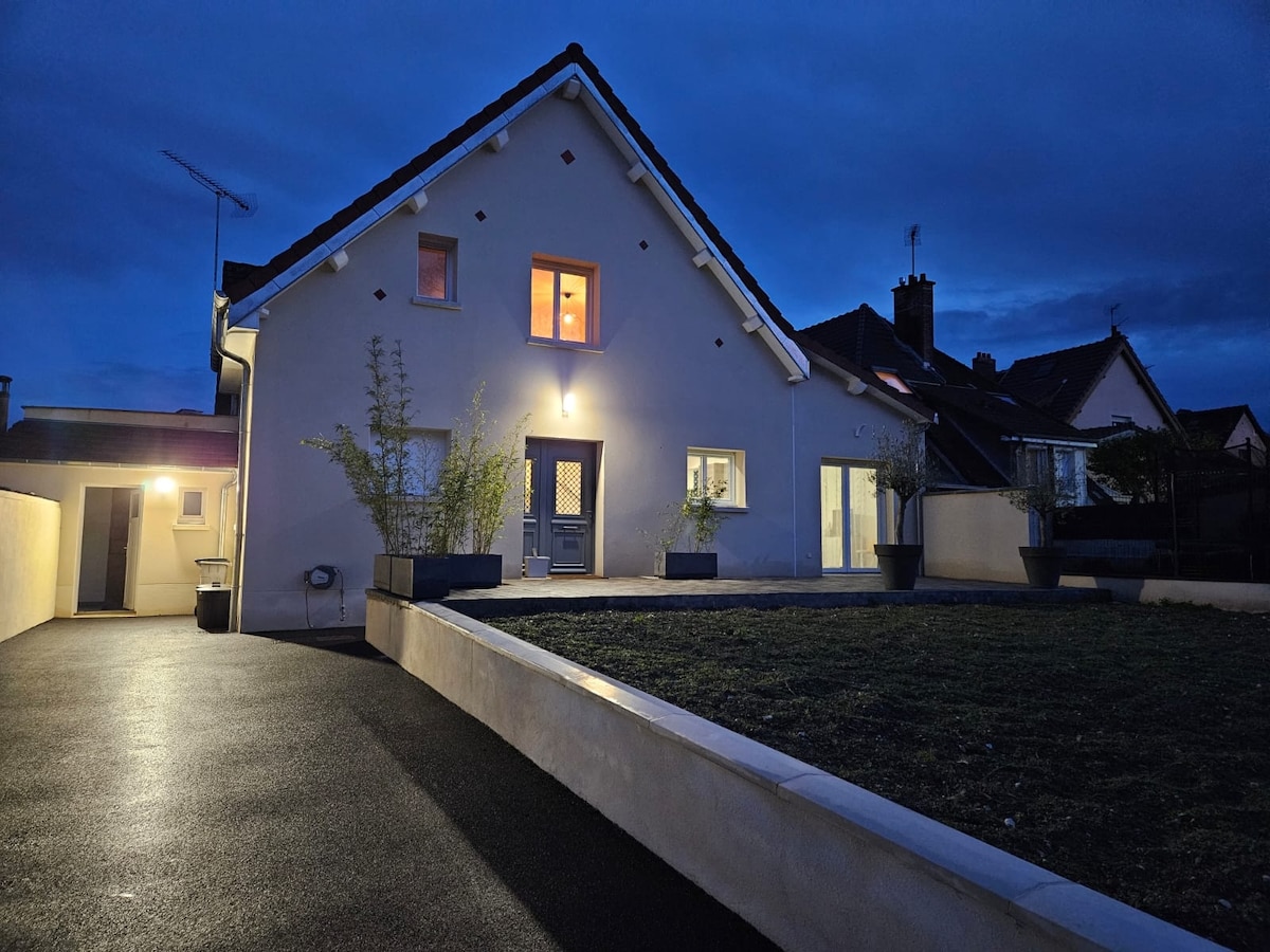 The exterior of the house is illuminated at dusk, highlighting its modern architecture with a sloped roof. Large windows provide a glimpse of the warm interior glow. Planters with greenery frame the entrance, and the driveway is spacious, leading to an open area of lawn.