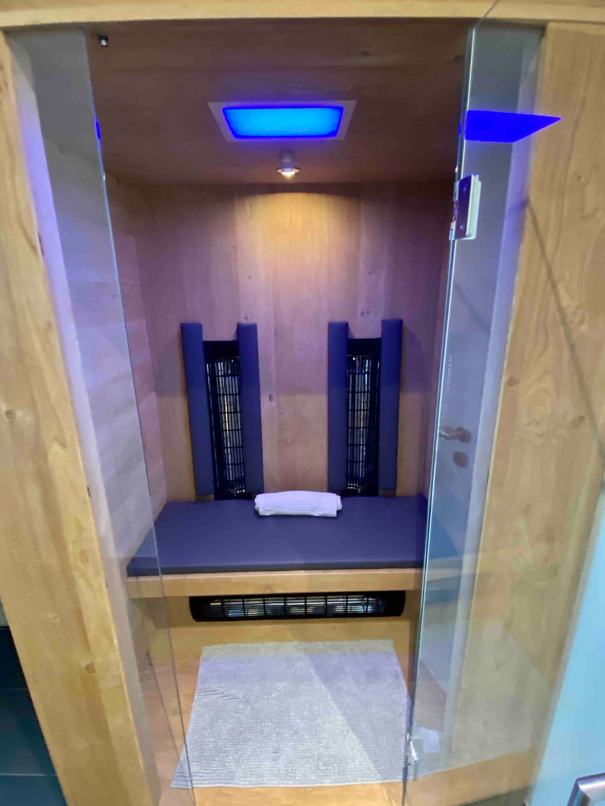 An infrared sauna is presented, featuring wooden interior walls and two black heating panels positioned on either side. A gray bench is situated inside with a white towel neatly placed on top. Soft blue lighting creates a calming atmosphere.