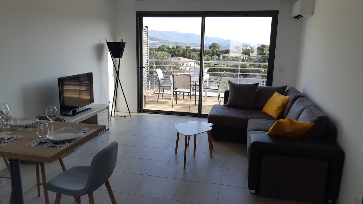 A modern living area features a comfortably arranged sectional sofa adorned with a yellow cushion. A small coffee table sits in front of a dining table set for two. Large glass doors provide access to a balcony with outdoor seating, offering a view of the surroundings.