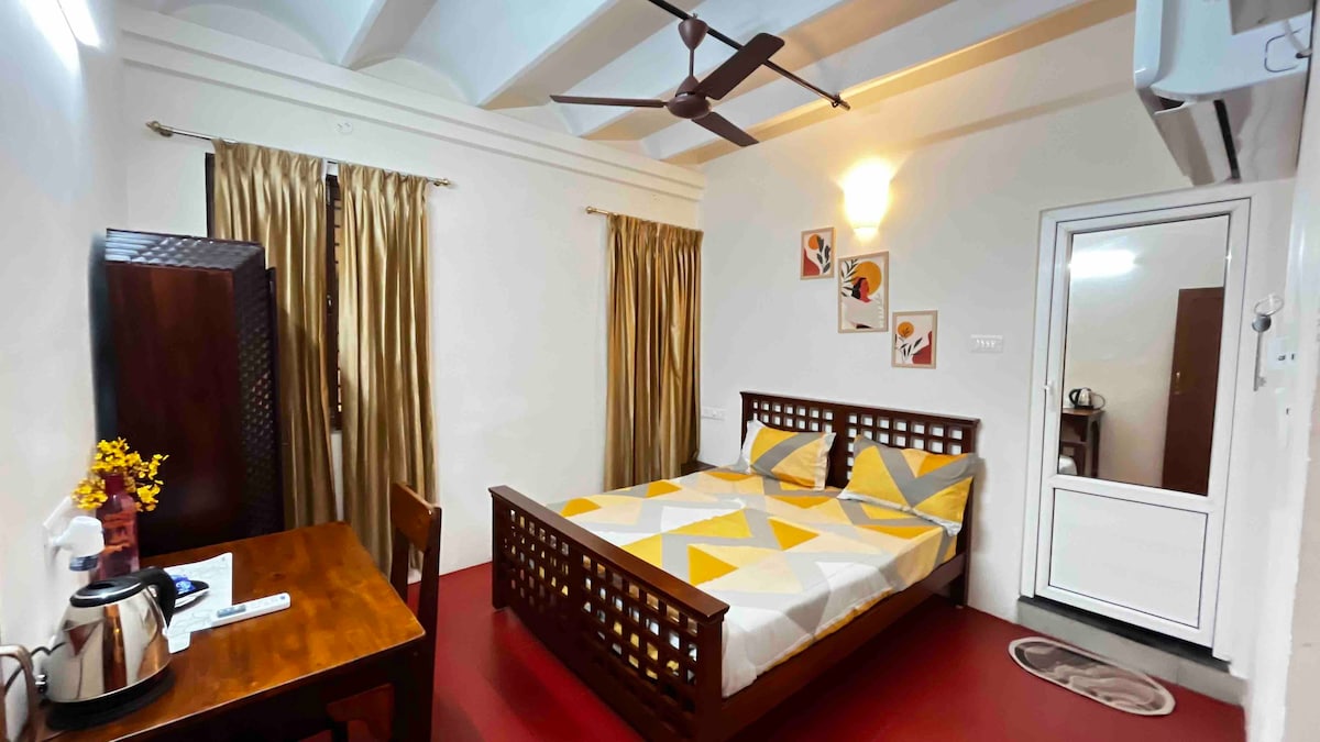 A well-furnished room features a spacious bed adorned with geometric-patterned linens. A wooden table is positioned nearby, accompanied by an electric kettle. Curtains frame the windows, blending with the room's warm tones. A private entrance to the washroom is visible in the background.