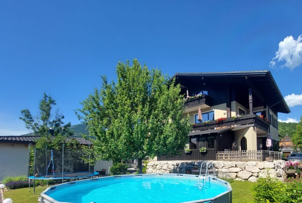 A generous outdoor space features a round pool surrounded by lush greenery. A large tree provides shade nearby, while the apartment house is visible in the background, showcasing its balconies adorned with flowers and clear blue skies overhead.
