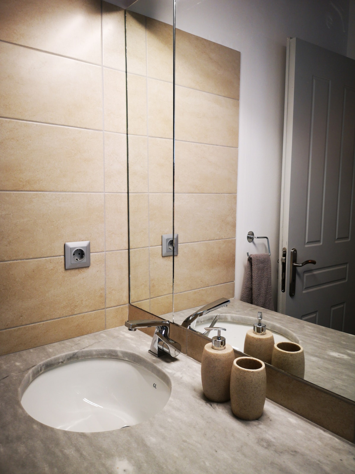 A modern bathroom featuring a stone countertop with a circular sink and a sleek faucet. A large mirror is mounted above the sink, reflecting warm-toned tiles on the wall. Decorative items, including two vases, are neatly arranged on the countertop.