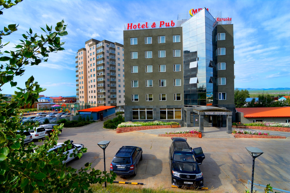 The exterior of MBM Hotel features a modern design with a glass front and the hotel’s name displayed prominently. Surrounding greenery and flower beds enhance the entrance area. Parking spaces are visible in front, with several vehicles parked along the hotel's facade.