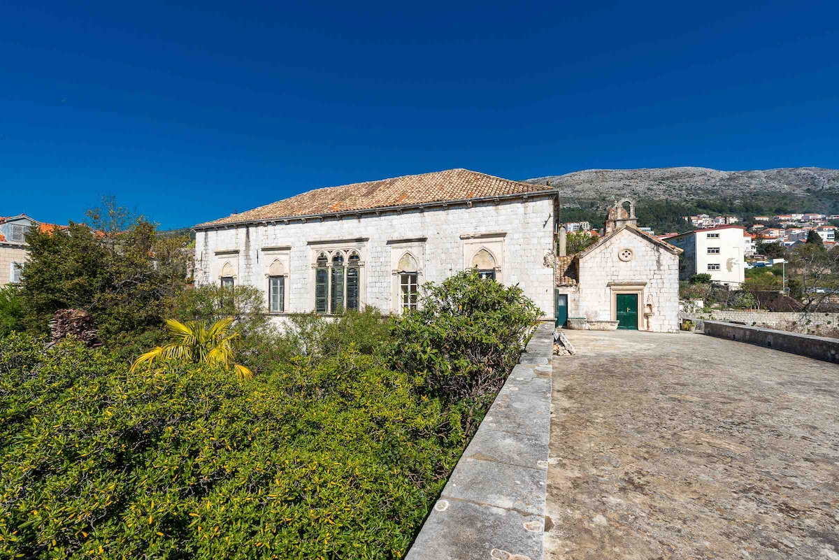 A historic stone building is featured, surrounded by greenery and set against a clear blue sky. The structure showcases arched windows and a traditional tiled roof, while a paved area in the foreground provides space for outdoor activities.