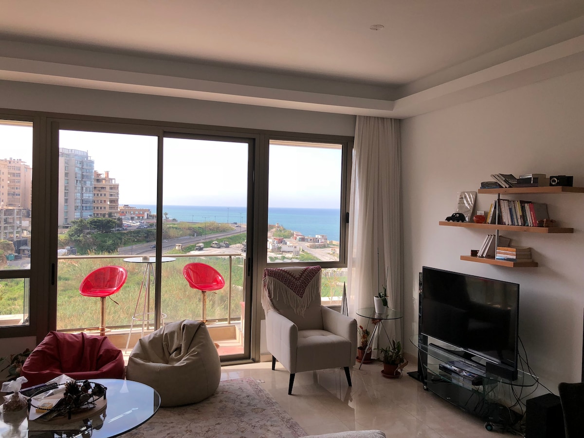 A modern living area is furnished with a comfortable chair and a seating pouf. A television set stands on a low unit, while a stylish shelf displays books. Large windows offer an unobstructed view of the Mediterranean Sea and the surrounding landscape.
