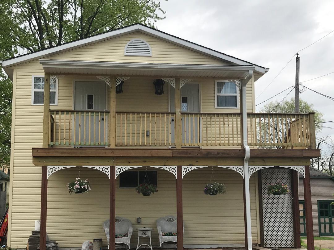 The exterior of a two-story house is presented, featuring a welcoming porch with wooden railings and hanging flower baskets. The light yellow exterior is complemented by white trim. A shaded area below includes comfortable seating, creating an inviting entrance to the upper-level apartments.