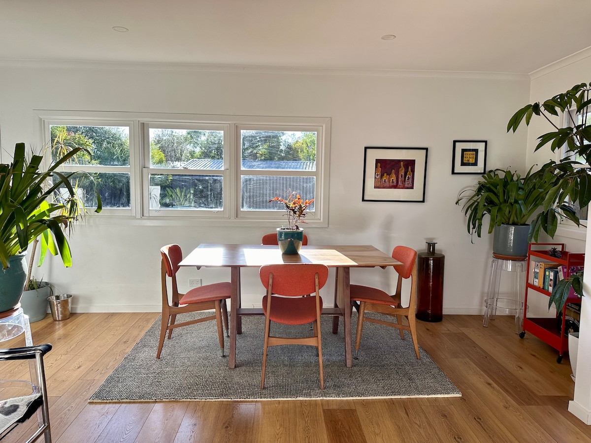 A dining area features a light wood table surrounded by four chairs, positioned on a soft rug. Large windows fill the space with natural light, highlighting greenery from potted plants. Art pieces are displayed on the walls, contributing to the home's vibrant decor.