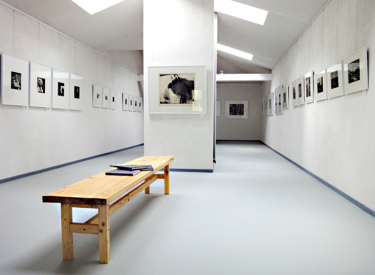 A spacious gallery is displayed, featuring white walls adorned with framed black-and-white photographs. A wooden bench occupies the center, providing a place for visitors to sit. Soft natural light floods the space through skylights, enhancing the minimalist aesthetic of the room.