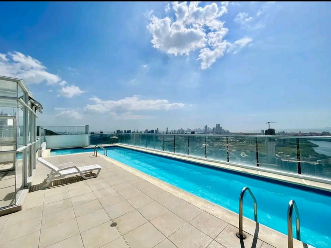A rooftop pool area is displayed, featuring a long, clear swimming pool surrounded by tiled flooring. Lounge chairs are positioned nearby, with a panoramic view of the city skyline and open sky observed in the background. Glass railing enhances safety while not obstructing the scenic view.