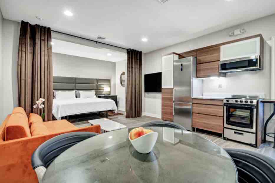 The studio features a modern interior with a spacious bed framed by elegant drapery. A sleek kitchen with stainless steel appliances and a dining table is visible, complemented by an orange sofa and a decorative bowl of apples on the table.