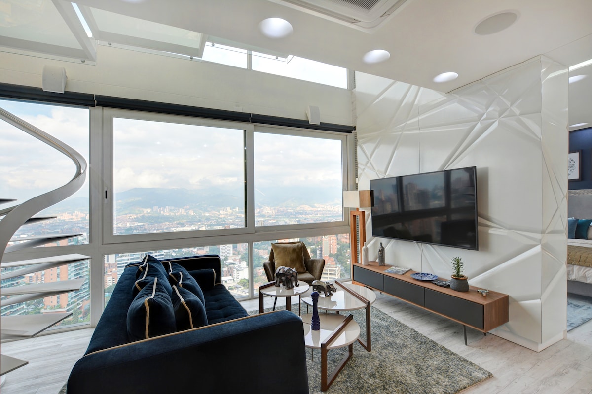 A modern living area features large windows that provide expansive views of the city valley. A dark blue sofa is accompanied by a light wood coffee table, while a sleek television is mounted on a textured wall. The spiral staircase adds architectural interest to the space.