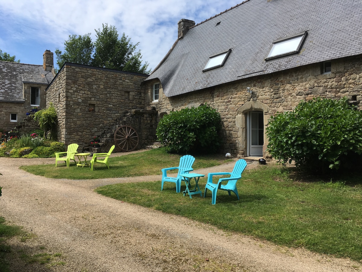 A gravel pathway meanders through a well-maintained garden area featuring green grass and stone structures. Colorful Adirondack chairs are arranged around a small table, inviting relaxation. The rustic architecture of the buildings, made from local stone, reflects traditional Breton style under a partly cloudy sky.