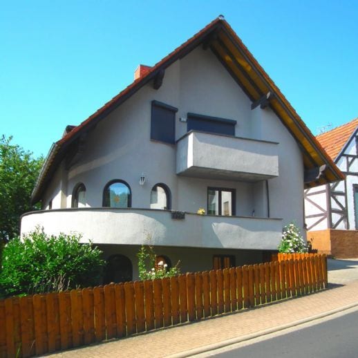 The exterior of a two-story house is displayed, featuring a unique architectural design with rounded windows and a spacious balcony. A wooden fence encloses a portion of the grassy area surrounding the home. The facade is complemented by surrounding greenery and a clear blue sky.
