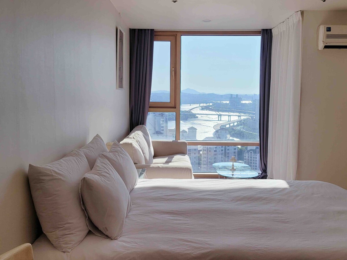 A spacious bed is positioned next to a large window, offering a scenic view of the Han River. Natural light filters through sheer curtains, creating a bright atmosphere. A small sofa and glass table complement the cozy setting, ideal for enjoying the surrounding scenery.