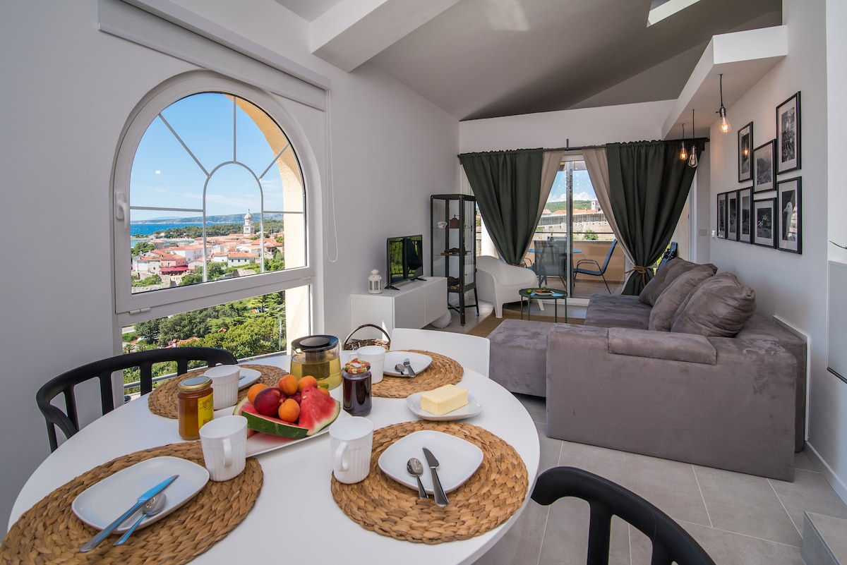 A bright dining area is presented with a round table set for breakfast, featuring plates, silverware, and a selection of fruit. The living space includes a comfortable gray sofa and a television, with large windows offering views of the surrounding landscape.
