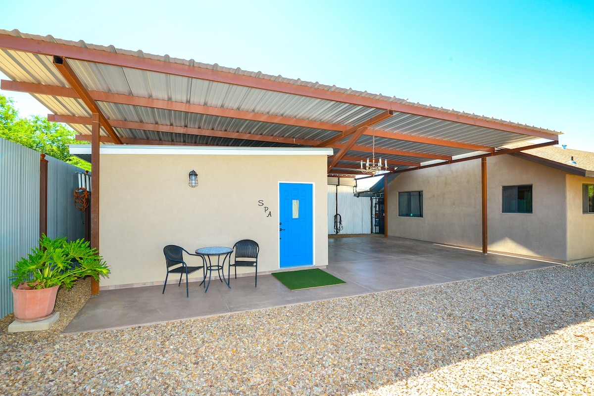 A covered outdoor patio area features a small round table with two chairs, situated on a gravel surface. The light-colored wall is complemented by a bold blue door with a decorative glass panel. A potted plant adds a touch of greenery to the scene.