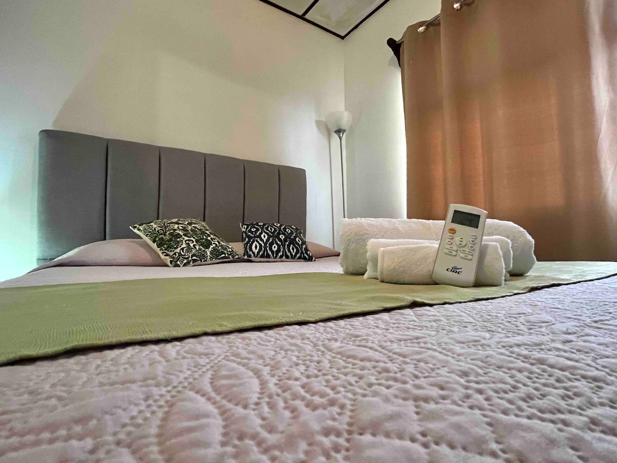 A spacious bed is showcased with a light green throw blanket and decorative cushions. Soft lighting from a nearby lamp creates a calming environment. Fresh towels are neatly arranged on the bed, complementing the pastel color palette of the room.