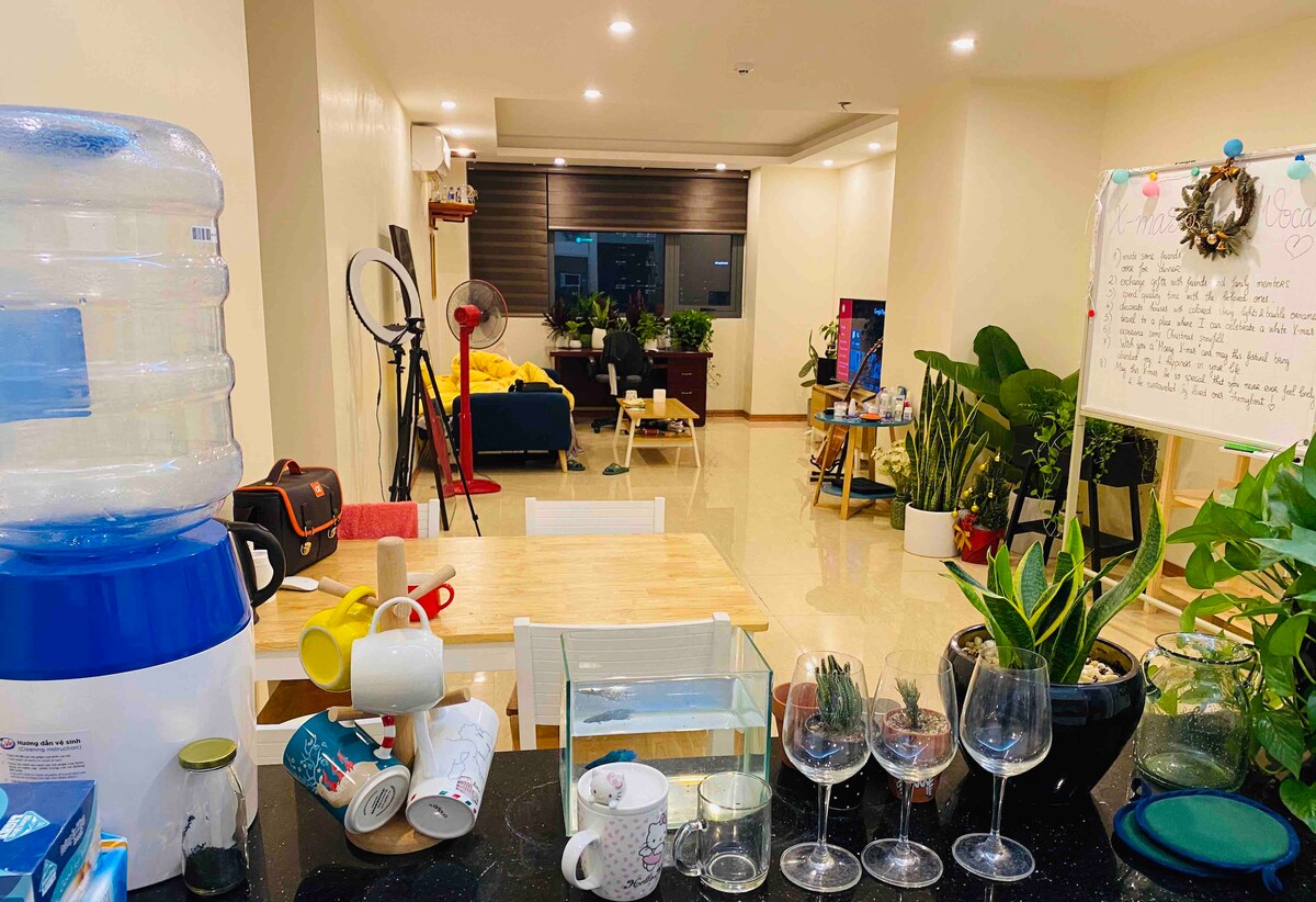 A shared living space is visible, featuring various plants arranged throughout, enhancing the green decor theme. A dining table is set with mugs and glassware. In the background, a cozy seating area awaits with a television, and natural light enters through large windows.