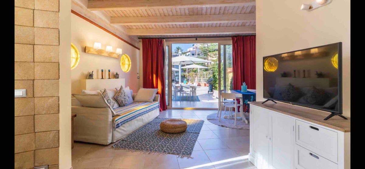 A bright living area is equipped with a comfortable sofa, accented by colorful pillows. Natural light fills the space through large sliding doors that open to a terrace with outdoor seating. A small dining table is positioned beside a modern television unit.