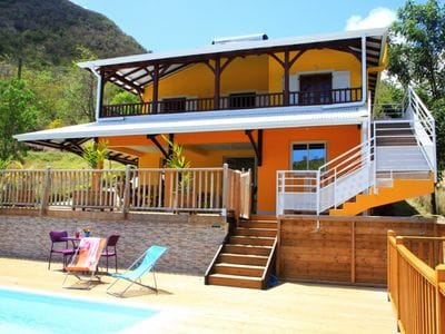 A two-story villa with a vibrant yellow exterior is surrounded by greenery. A wooden deck leads to a spacious pool area, featuring lounge chairs and a table for relaxation. A staircase provides access to the upper level, where large windows invite natural light.