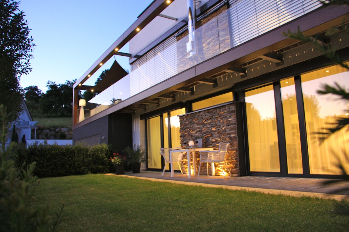 A modern exterior view of the apartment showcases large glass doors that open to a private terrace. Comfortable outdoor seating is positioned on a manicured lawn, framed by neatly trimmed hedges. Soft outdoor lighting highlights the building's contemporary design against the twilight sky.