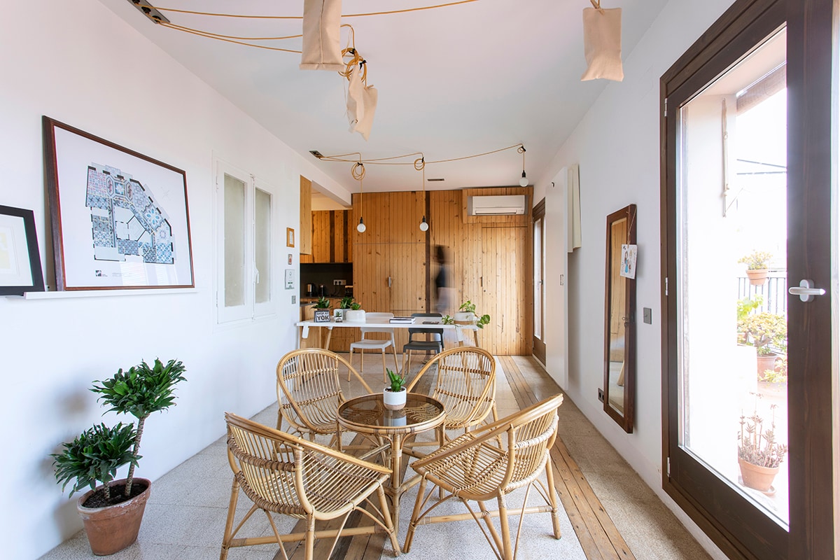 A modern living area is featured, showcasing a round table surrounded by four rattan chairs. Natural light pours in through large glass doors, highlighting the eco-friendly decor. A kitchen area is visible in the background, with wooden accents and minimalistic design elements.