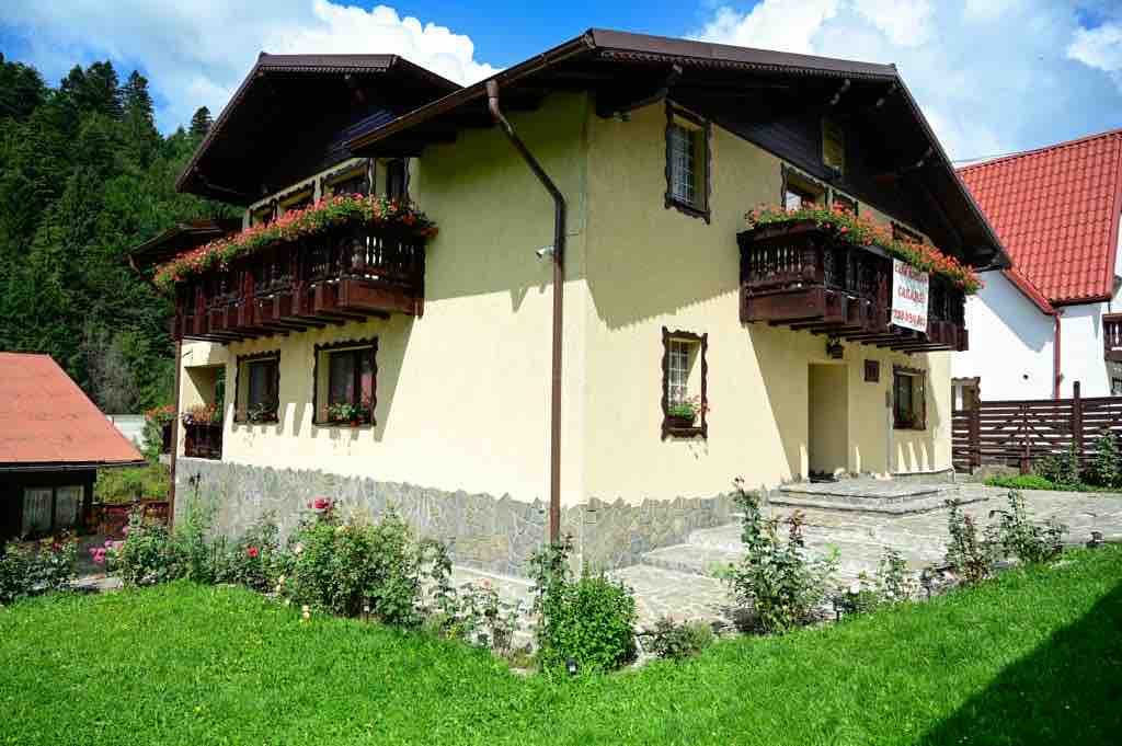 The exterior of a two-story building showcases a yellow facade with wooden balconies adorned with vibrant flowers. A stone pathway leads through well-maintained green grass, bordered by blooming rose bushes. A contrasting red-roofed structure is visible in the background, surrounded by trees.