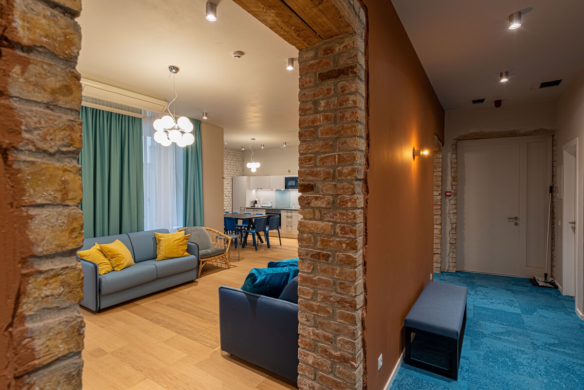 A spacious hallway leads to a well-lit living area featuring a blue sofa with decorative pillows. Soft curtains frame the windows, while an adjacent kitchen area is visible in the background. Exposed brick walls add texture to the warm-toned environment.