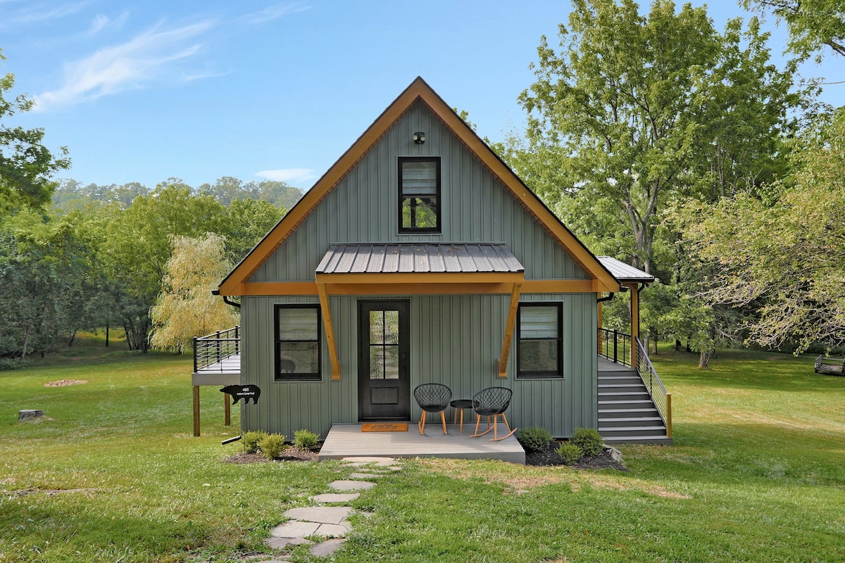 The cabin features a welcoming exterior with a green facade and wooden accents. A set of steps leads to the front door, flanked by two black chairs. A grill is positioned nearby, and lush greenery surrounds the property, contributing to a serene outdoor setting.