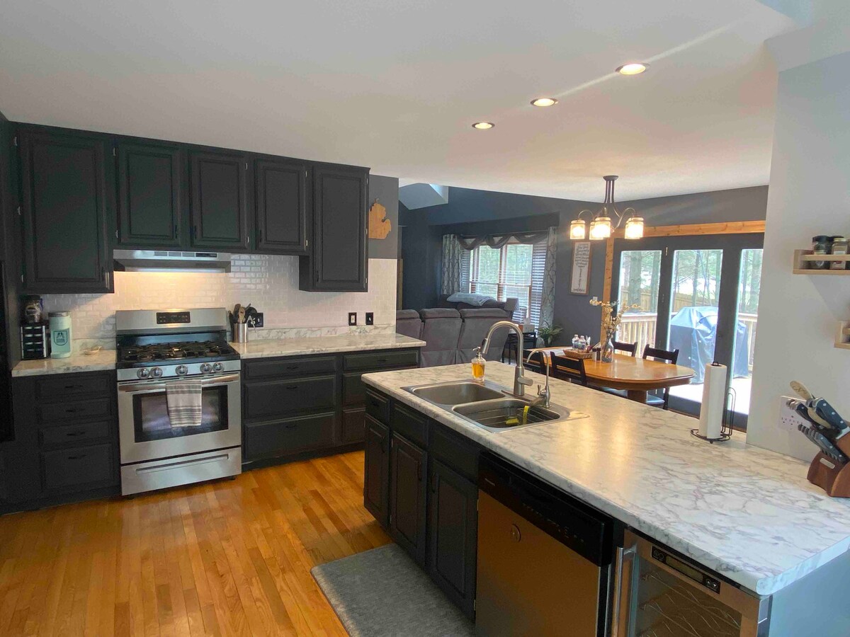 The kitchen features dark cabinets and marble countertops, illuminated by overhead lights. A stainless-steel gas range is present alongside a double sink. The space leads to a dining area with large windows overlooking the backyard, complemented by a cozy seating arrangement.