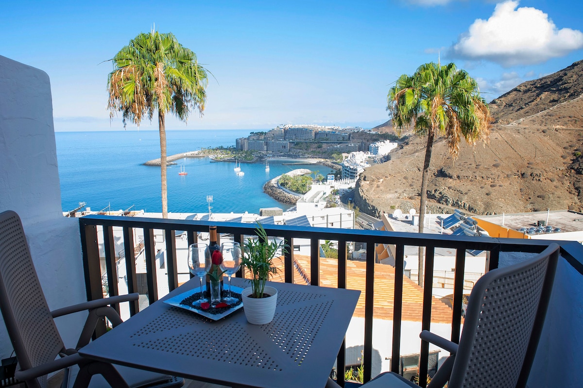 A private balcony showcases a scenic view of the coastline and mountains beyond. Two chairs and a small table are present, adorned with a decorative tray featuring two glasses and a potted plant. Palm trees are visible, contributing to the serene atmosphere.