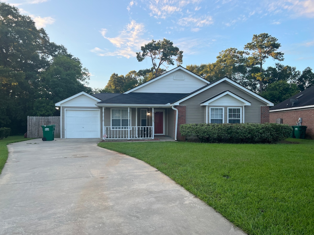 The front exterior of the home features a well-maintained yard with grass and landscaped bushes. A paved driveway leads to a garage and a covered porch area. Trees are visible in the background, providing natural shade.