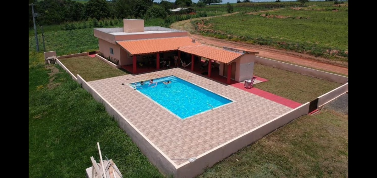An aerial view of a spacious outdoor area features a rectangular pool surrounded by a tiled deck. A red-paved patio offers additional space for relaxation. Lush green fields are visible in the background, enhancing the scene's open and airy feel.
