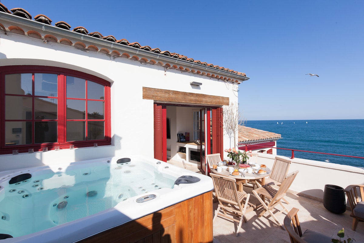 A spacious terrace featuring a hot tub, surrounded by wooden deck chairs and a dining table. Large red-framed windows open to reveal a view of the sea, enhancing the tranquil ambiance. The clear blue sky complements the picturesque coastal scenery.