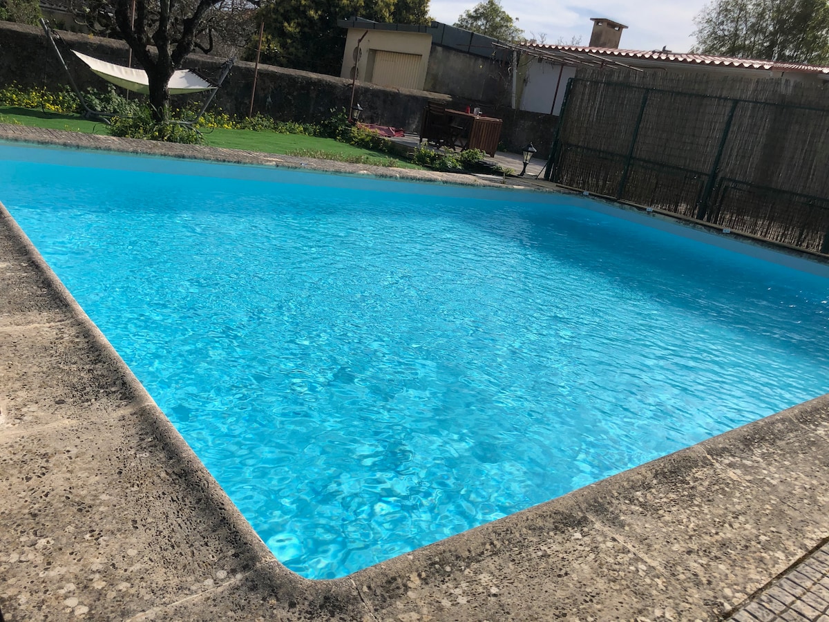 A sparkling blue pool reflects the sunlight, surrounded by a concrete deck. Lush greenery and a hammock are visible in the background, offering a peaceful outdoor setting. The fence provides a sense of privacy, enhancing the tranquil atmosphere.