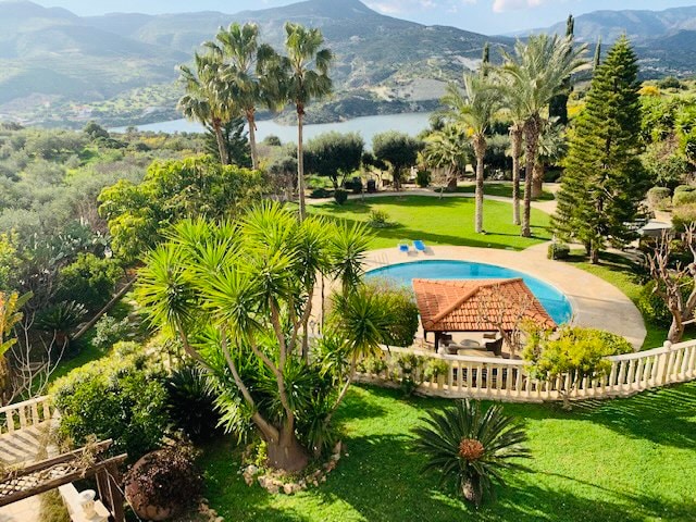 A lush garden area features a swimming pool surrounded by palm trees and other greenery, overlooking Germasoyia Dam. A shaded outdoor seating area is visible near the pool, complemented by well-maintained lawns and a backdrop of rolling mountains.