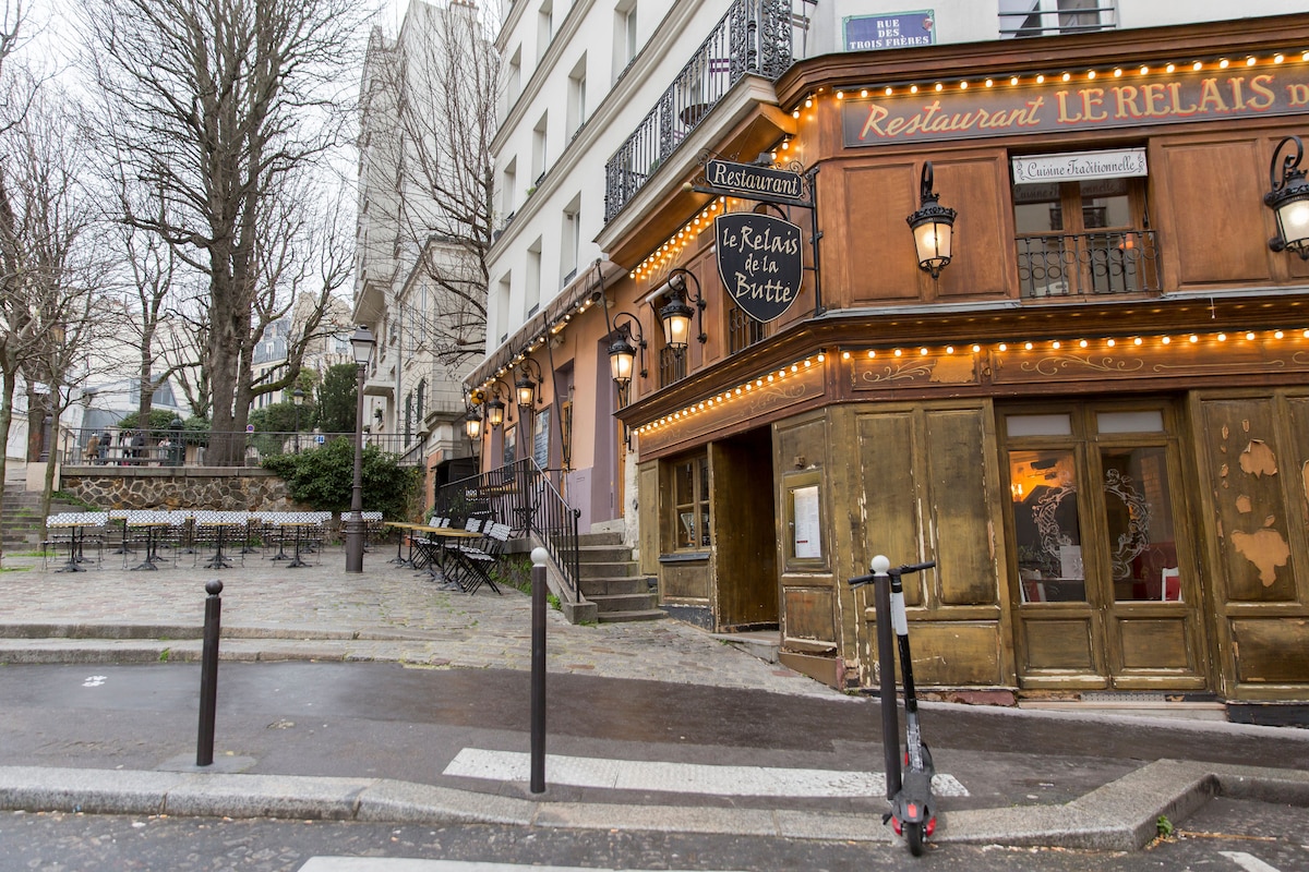 A charming restaurant exterior is seen, featuring ornate wooden details and string lights. Outdoor seating is available with tables and chairs set against a backdrop of trees. A nearby scooter is positioned on the pavement, adding to the vibrant street scene.