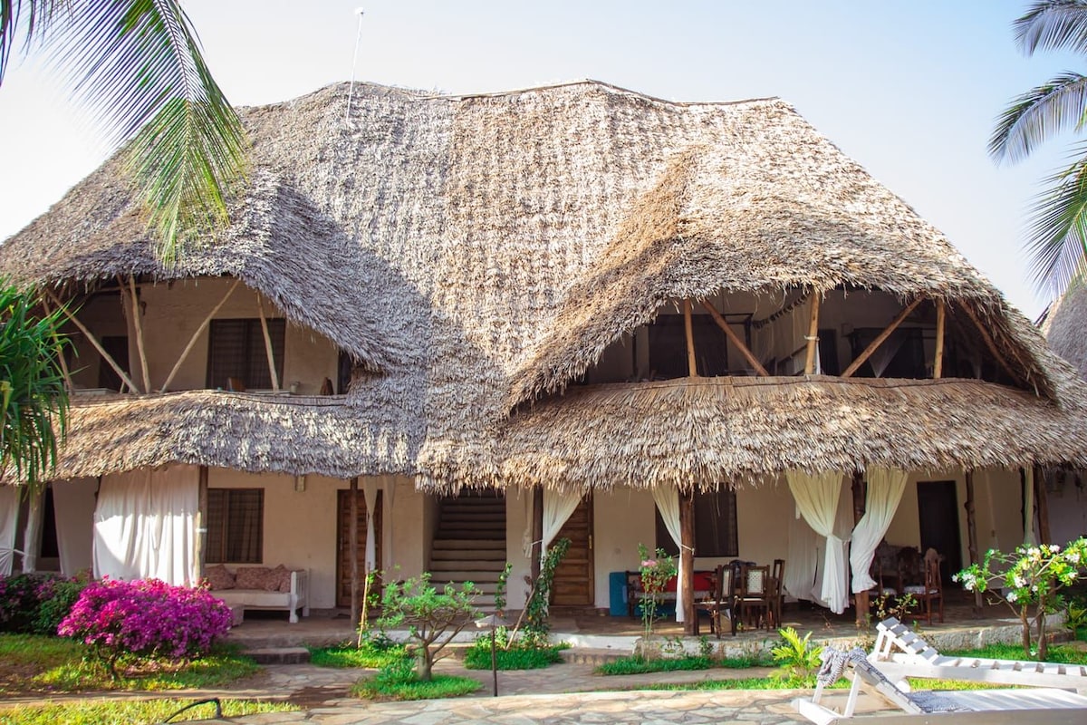 A charming two-story building features a thatched roof and open verandas, surrounded by lush greenery and vibrant bougainvillea. The ground level includes a seating area and dining space, with large windows providing natural light to the interior.