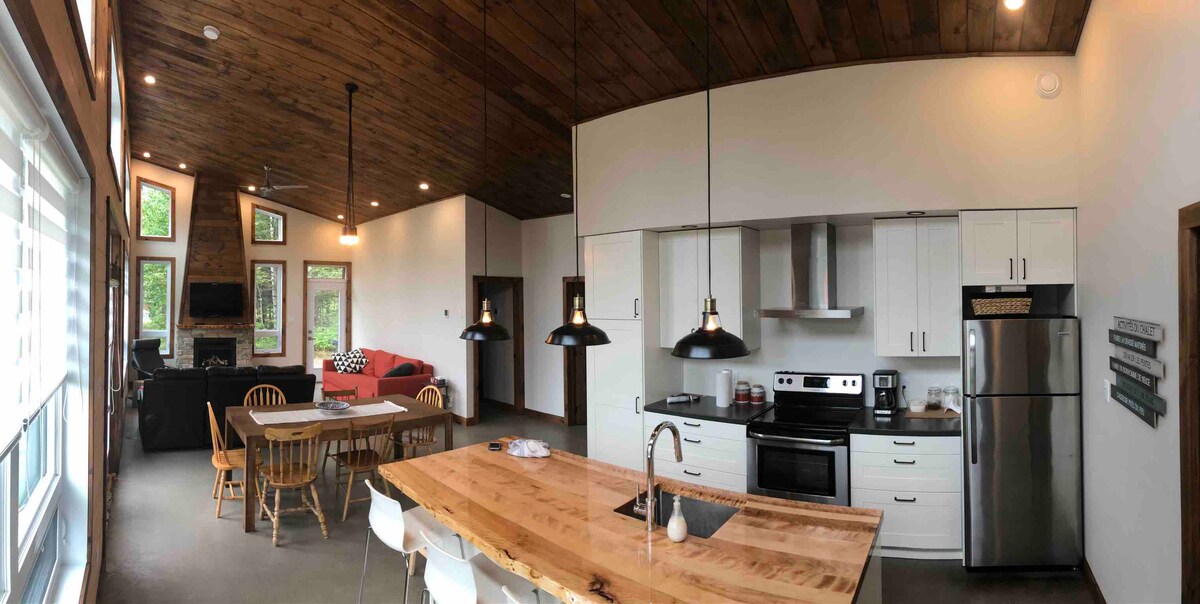 A modern kitchen area features a spacious wooden island with seating. Black pendant lights hang above, illuminating the countertop. The kitchen is equipped with sleek appliances, and an adjacent dining table is visible. Large windows allow natural light to enter, enhancing the open layout.
