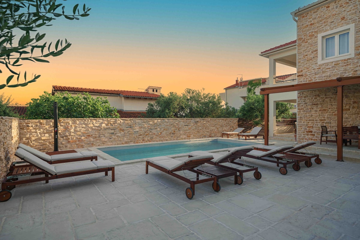 The outdoor area features a private pool surrounded by a stone wall, with several sun loungers positioned for relaxation. A patio is visible in the background, complemented by chairs and a table, all set against a backdrop of sunset hues.