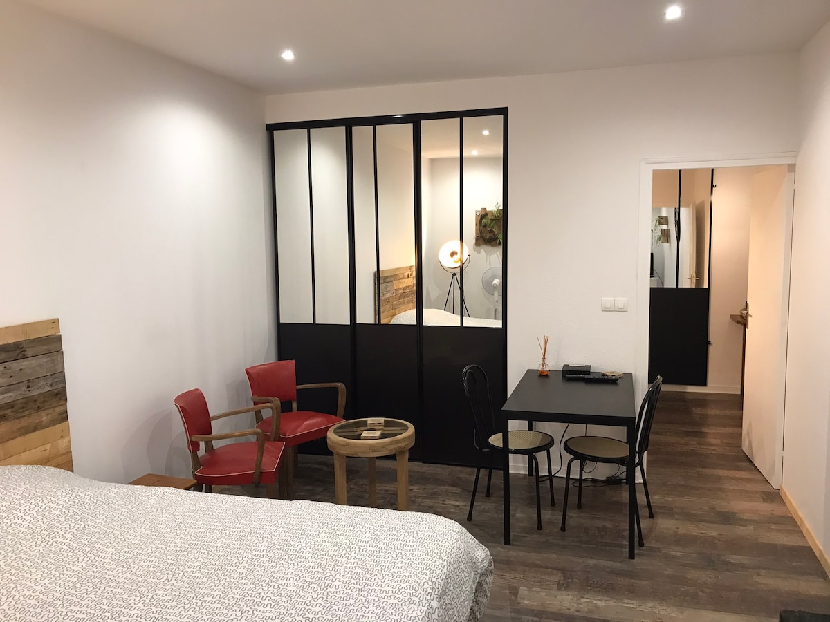 A cozy living area features a spacious bed with a textured light-colored cover. A glass partition separates the sleeping space from a small dining area, furnished with a round table and two chairs. Light fixtures and subtle wooden accents enhance the overall design.