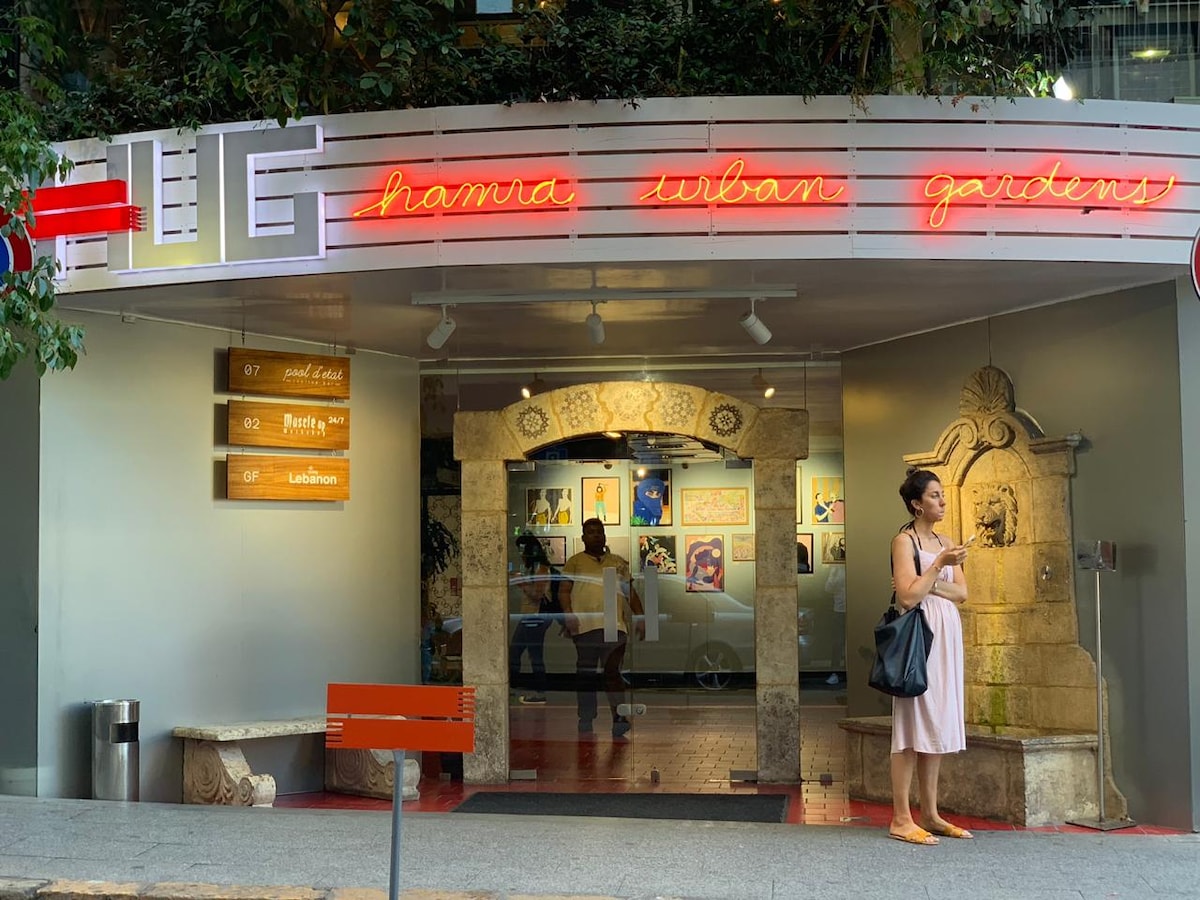 The entrance of HUG is highlighted by a vibrant neon sign featuring the name, framed by lush greenery. A stone archway leads into the interior, where art pieces are displayed. Directional signs provide information about various facilities nearby.